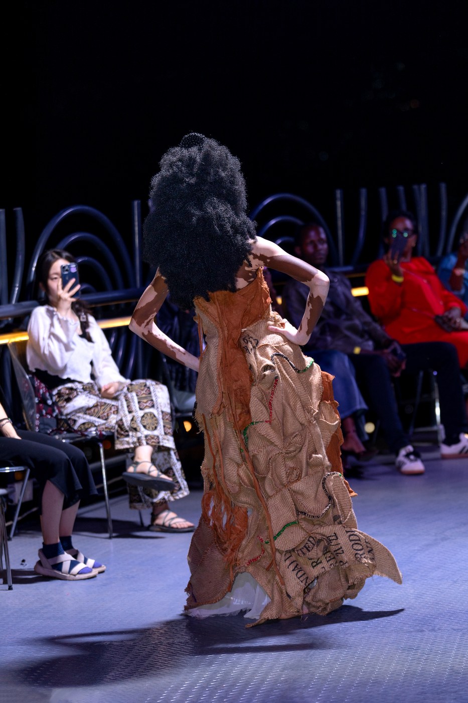 Back view of model in voluminous gunia sack dress with original market print lettering visible on the fabric — Bobbin Case runway Kigali 2026
